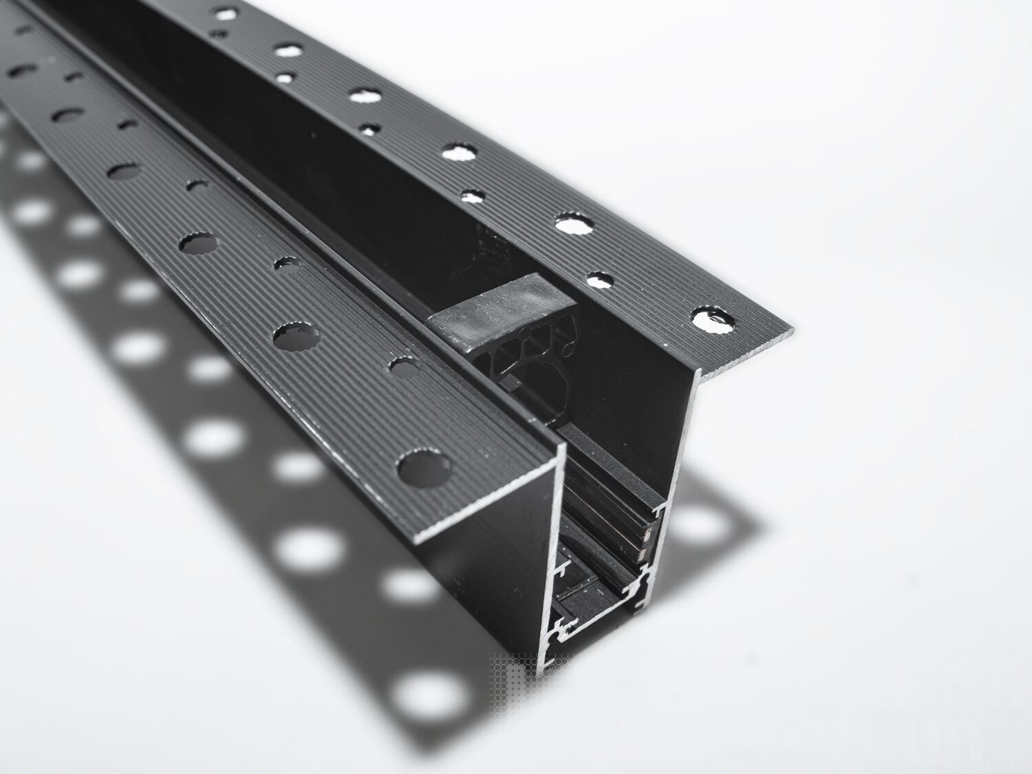 Рейлінг-шинопровід для магнітної трекової системи, розроблений для прихованої установки, є сучасним і технологічно вдосконаленим рішенням для створення гнучких освітлювальних сценаріїв. Ця система освітлення, що ледь помітна на стелі або стіні, ідеально підходить для тих, хто прагне до мінімалістичного та витонченого дизайну.
Шинопровід являє собою металевий профіль, який монтується врівень із поверхнею стелі або стіни, зазвичай перед покриттям гіпсокартоном або іншими оздоблювальними матеріалами. Він може бути встановлений між стельовими балками або на спеціальні монтажні планки, що забезпечують необхідну підтримку. Для монтажу цього треку достатньо штроби глибиною 85мм і шириною 45мм (у зовнішній частині 90 мм для фіксації шаром штукатурки закладних елементів). Після установки шинопроводу, поверхня стелі обробляється таким чином, що лише декоративна частина освітлювальних приладів залишається видимою.
Всередині шинопроводу проходять електричні проводи, які забезпечують живлення світильників. Магнітні компоненти в шинопроводі дозволяють легко прикріплювати або переміщувати світильники, тим самим, надаючи можливість змінювати освітлення відповідно до поточних потреб або перепланування простору.
Шинопровід складається із двох пар провідників. Одна пара для живлення світильників. Друга резервна для підключення управління світильниками, наприклад, за допомогою контролерів стандарту DALI.
Шинопровід розрахований на використання напруги живлення 48В. У конструкції використані шини прямокутного перерізу, які забезпечують більшу площу перерізу провідника, та кращий контакт, ніж шини з круглим перетином.
Додатковою перевагою є те, що така прихована система дозволяє уникнути видимого нагромадження технічних елементів на стелі, зберігаючи естетичну чистоту дизайну. Це створює ілюзію, що світло виходить прямо з поверхні, додаючи відчуття простору і візуально розширюючи кімнату.
Рейлінг-шинопровід для прихованої установки може мати різні форми і розміри, що надає додаткової гнучкості при проектуванні освітлювальних рішень. Він також може бути пофарбований у колір стелі, щоб ще більше інтегруватися в інтер'єр.
Завдяки використанню стандартного профілю для системи 48В, можна комбінувати треки та світильники різних виробників в одній системі.
Такі системи часто застосовуються в комерційних і житлових просторах, які підкреслюють сучасний дизайн інтер'єру, де освітлення є ключовим елементом декору. Вони забезпечують не тільки функціональне освітлення, а й виконують роль декоративного елементу, додаючи інтер'єру завершеності та стилю.