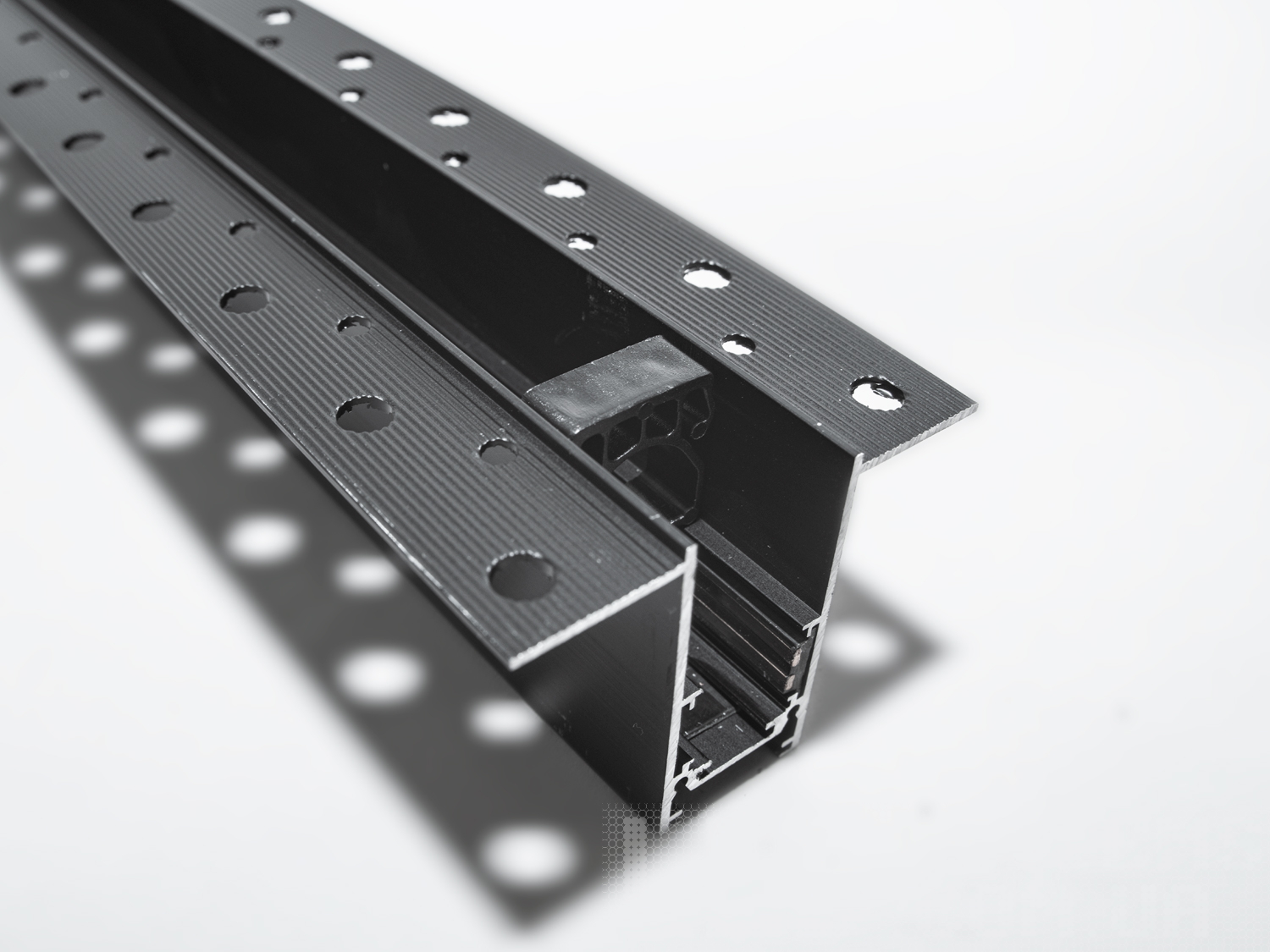 Рейлінг-шинопровід для магнітної трекової системи, розроблений для прихованої установки, є сучасним і технологічно вдосконаленим рішенням для створення гнучких освітлювальних сценаріїв. Ця система освітлення, що ледь помітна на стелі або стіні, ідеально підходить для тих, хто прагне до мінімалістичного та витонченого дизайну.
Шинопровід являє собою металевий профіль, який монтується врівень із поверхнею стелі або стіни, зазвичай перед покриттям гіпсокартоном або іншими оздоблювальними матеріалами. Він може бути встановлений між стельовими балками або на спеціальні монтажні планки, що забезпечують необхідну підтримку. Для монтажу цього треку достатньо штроби глибиною 85мм і шириною 45мм (у зовнішній частині 90 мм для фіксації шаром штукатурки закладних елементів). Після установки шинопроводу, поверхня стелі обробляється таким чином, що лише декоративна частина освітлювальних приладів залишається видимою.
Всередині шинопроводу проходять електричні проводи, які забезпечують живлення світильників. Магнітні компоненти в шинопроводі дозволяють легко прикріплювати або переміщувати світильники, тим самим, надаючи можливість змінювати освітлення відповідно до поточних потреб або перепланування простору.
Шинопровід складається із двох пар провідників. Одна пара для живлення світильників. Друга резервна для підключення управління світильниками, наприклад, за допомогою контролерів стандарту DALI.
Шинопровід розрахований на використання напруги живлення 48В. У конструкції використані шини прямокутного перерізу, які забезпечують більшу площу перерізу провідника, та кращий контакт, ніж шини з круглим перетином.
Додатковою перевагою є те, що така прихована система дозволяє уникнути видимого нагромадження технічних елементів на стелі, зберігаючи естетичну чистоту дизайну. Це створює ілюзію, що світло виходить прямо з поверхні, додаючи відчуття простору і візуально розширюючи кімнату.
Рейлінг-шинопровід для прихованої установки може мати різні форми і розміри, що надає додаткової гнучкості при проектуванні освітлювальних рішень. Він також може бути пофарбований у колір стелі, щоб ще більше інтегруватися в інтер'єр.
Завдяки використанню стандартного профілю для системи 48В, можна комбінувати треки та світильники різних виробників в одній системі.
Такі системи часто застосовуються в комерційних і житлових просторах, які підкреслюють сучасний дизайн інтер'єру, де освітлення є ключовим елементом декору. Вони забезпечують не тільки функціональне освітлення, а й виконують роль декоративного елементу, додаючи інтер'єру завершеності та стилю.