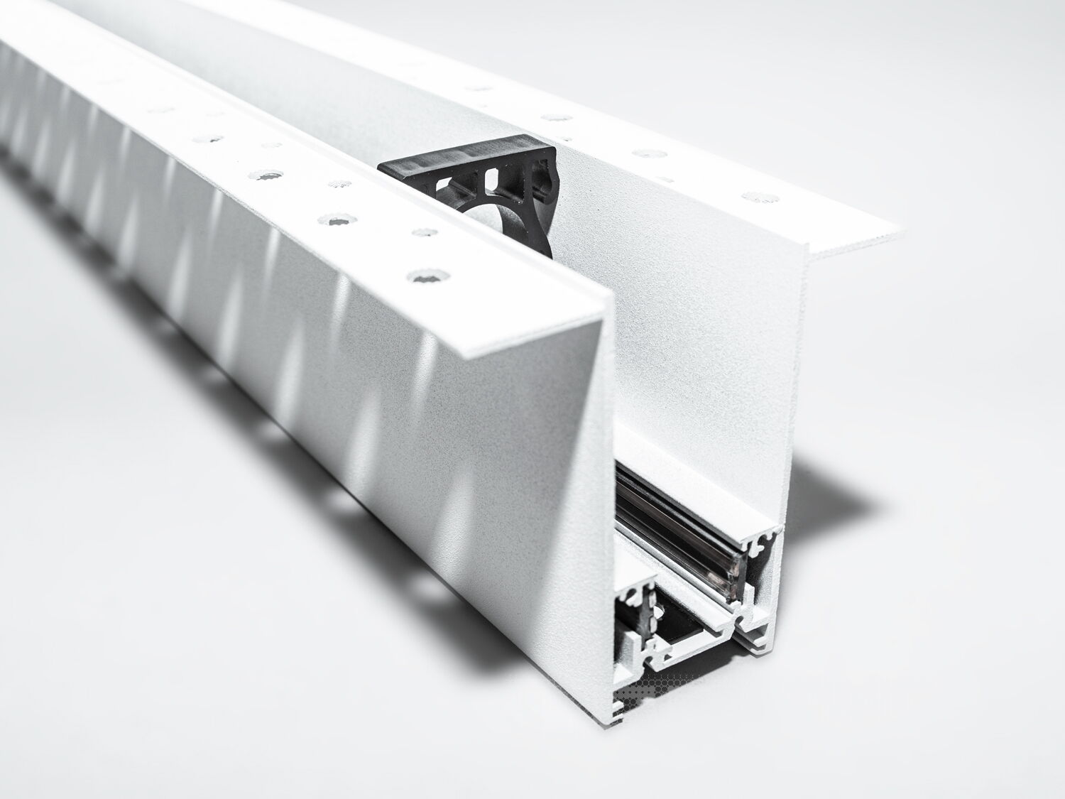 Рейлінг-шинопровід для магнітної трекової системи, розроблений для прихованої установки, є сучасним і технологічно вдосконаленим рішенням для створення гнучких освітлювальних сценаріїв. Ця система освітлення, що ледь помітна на стелі або стіні, ідеально підходить для тих, хто прагне до мінімалістичного та витонченого дизайну.
Шинопровід являє собою металевий профіль, який монтується врівень із поверхнею стелі або стіни, зазвичай перед покриттям гіпсокартоном або іншими оздоблювальними матеріалами. Він може бути встановлений між стельовими балками або на спеціальні монтажні планки, що забезпечують необхідну підтримку. Для монтажу цього треку достатньо штроби глибиною 85мм і шириною 45мм (у зовнішній частині 90 мм для фіксації шаром штукатурки закладних елементів). Після установки шинопроводу, поверхня стелі обробляється таким чином, що лише декоративна частина освітлювальних приладів залишається видимою.
Всередині шинопроводу проходять електричні проводи, які забезпечують живлення світильників. Магнітні компоненти в шинопроводі дозволяють легко прикріплювати або переміщувати світильники, тим самим, надаючи можливість змінювати освітлення відповідно до поточних потреб або перепланування простору.
Шинопровід складається із двох пар провідників. Одна пара для живлення світильників. Друга резервна для підключення управління світильниками, наприклад, за допомогою контролерів стандарту DALI.
Шинопровід розрахований на використання напруги живлення 48В. У конструкції використані шини прямокутного перерізу, які забезпечують більшу площу перерізу провідника, та кращий контакт, ніж шини з круглим перетином.
Додатковою перевагою є те, що така прихована система дозволяє уникнути видимого нагромадження технічних елементів на стелі, зберігаючи естетичну чистоту дизайну. Це створює ілюзію, що світло виходить прямо з поверхні, додаючи відчуття простору і візуально розширюючи кімнату.
Рейлінг-шинопровід для прихованої установки може мати різні форми і розміри, що надає додаткової гнучкості при проектуванні освітлювальних рішень. Він також може бути пофарбований у колір стелі, щоб ще більше інтегруватися в інтер'єр.
Завдяки використанню стандартного профілю для системи 48В, можна комбінувати треки та світильники різних виробників в одній системі.
Такі системи часто застосовуються в комерційних і житлових просторах, які підкреслюють сучасний дизайн інтер'єру, де освітлення є ключовим елементом декору. Вони забезпечують не тільки функціональне освітлення, а й виконують роль декоративного елементу, додаючи інтер'єру завершеності та стилю.