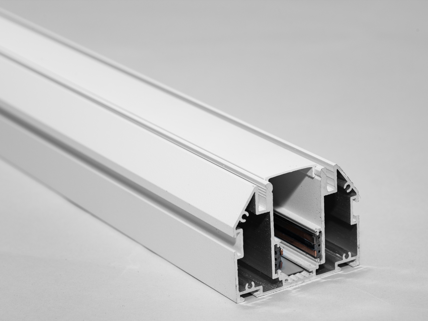 Рейлінг-шинопровід для магнітної трекової системи, призначений для натяжної стелі, представляє собою практичне та інноваційне рішення для сучасного освітлення. Цей тип шинопроводу спеціально розроблений для інтеграції з натяжними стельовими системами, які використовуються для створення гладких та візуально вільних поверхонь.
Шинопровід інсталюється безпосередньо на несучу основу стелі або на спеціальну підвісну систему перед установкою самої натяжної плівки або тканини. Це забезпечує надійність конструкції та гарантує, що шинопровід залишиться невидимим після монтажу натяжної стелі. Рейлінг повинен мати адекватну несучу здатність для того, щоб витримувати вагу світильників, що будуть до нього прикріплені.
Шинопровід складається із двох пар провідників. Одна пара для живлення світильників. Друга резервна для підключення управління світильниками, наприклад, за допомогою контролерів стандарту DALI.
Шинопровід розрахований на використання напруги живлення 48В. У конструкції використані шини прямокутного перерізу, які забезпечують більшу площу перерізу провідника, та кращий контакт, ніж шини з круглим перетином.
Завдяки магнітній системі, світильники можна легко прикріплювати до шинопроводу і переміщувати по ньому, що дозволяє легко змінювати освітлювальні сценарії. Це особливо корисно в місцях, де часто потрібно оновлювати або змінювати освітлення, таких як комерційні простори, галереї або багатофункціональні зали.
Освітлювальна система для натяжних стель також має бути добре продуманою з точки зору теплового менеджменту. Світлодіодні світильники зазвичай виробляють менше тепла ніж традиційні освітлювальні прилади, але важливо врахувати вентиляцію та тепловідведення, щоб запобігти нагріванню натяжної плівки чи тканини.
Використання рейлінг-шинопроводу для натяжної стелі дозволяє створити ефект 